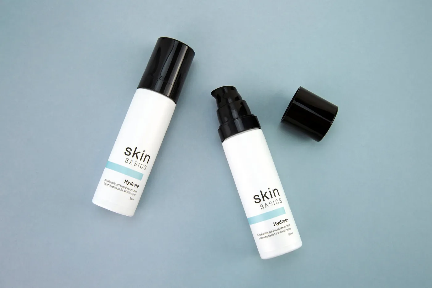 Two white airless bottles of a client's white label Hydrate serum with an aqua label band, lying on a pale blue surface